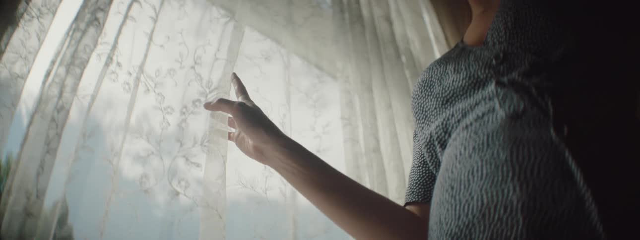 Woman with a blue dress touches curtains and looks out the window