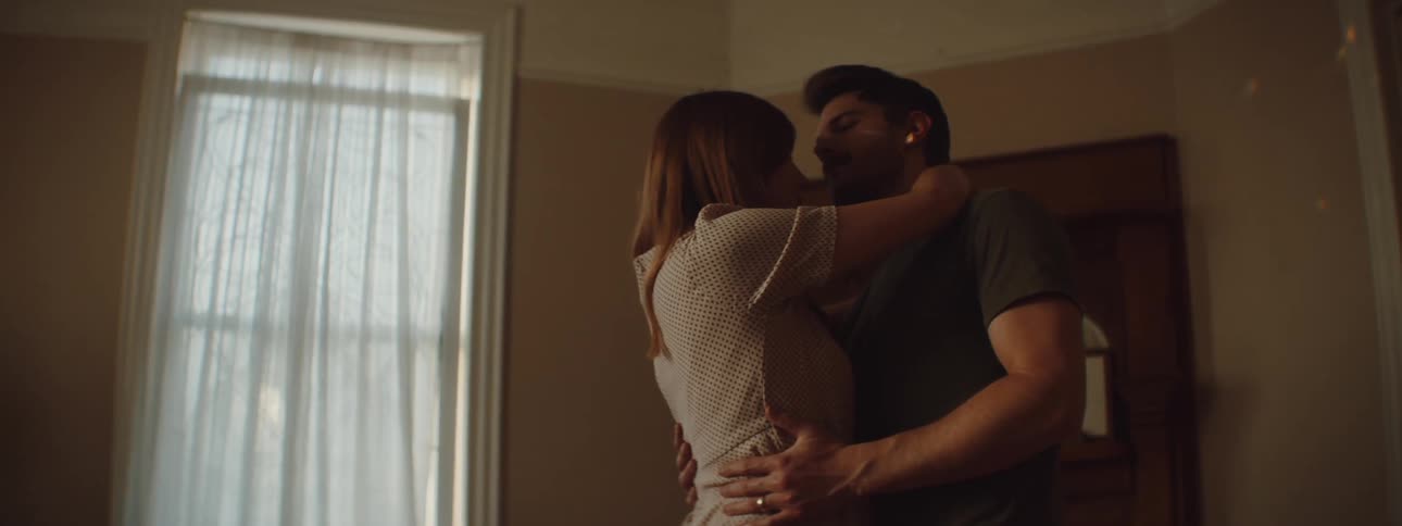 A man and a woman in a room slow dancing with a wall and window in the background