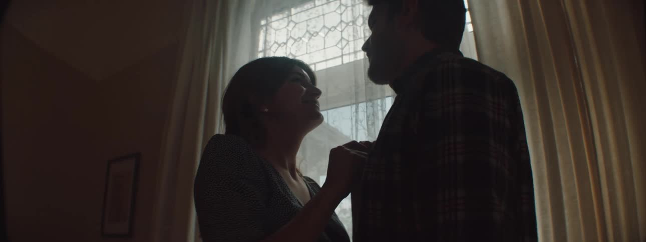 Husband and a pregnant wife playing around and poking at each other while standing in front of a window at home