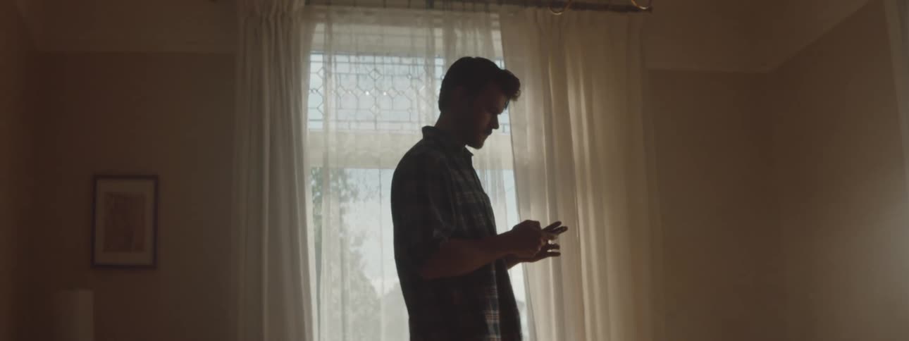 A man stands in a room indoors during the day while texting on a cellphone