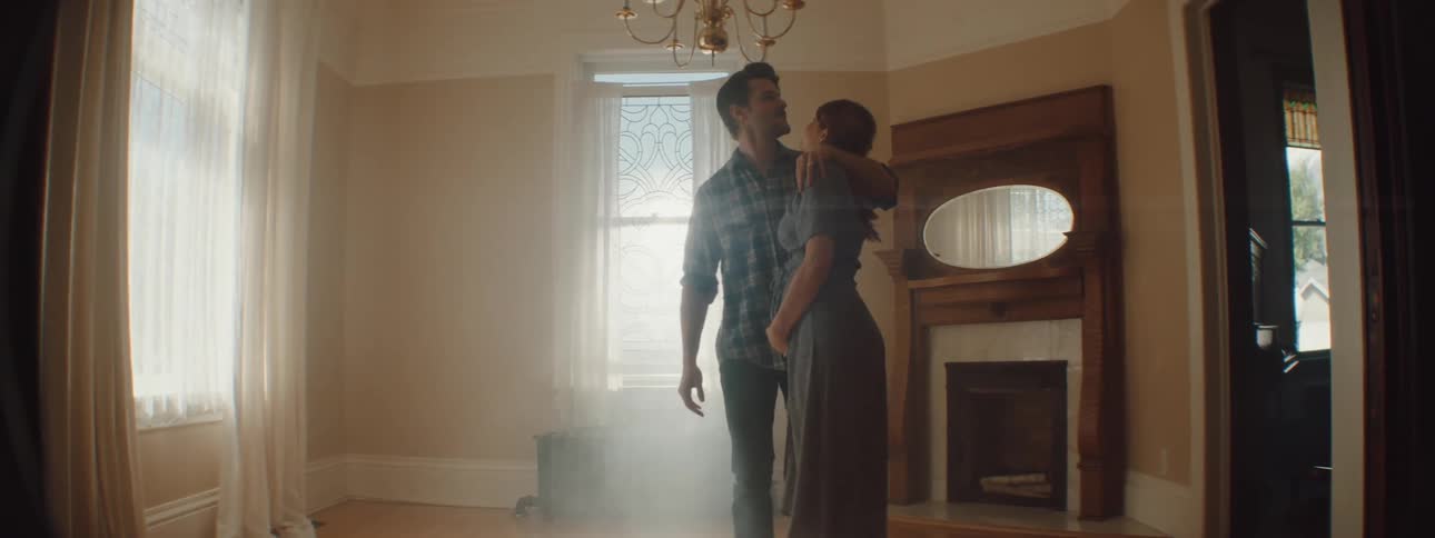 A man and woman holding hands walk in to an empty living room and hug