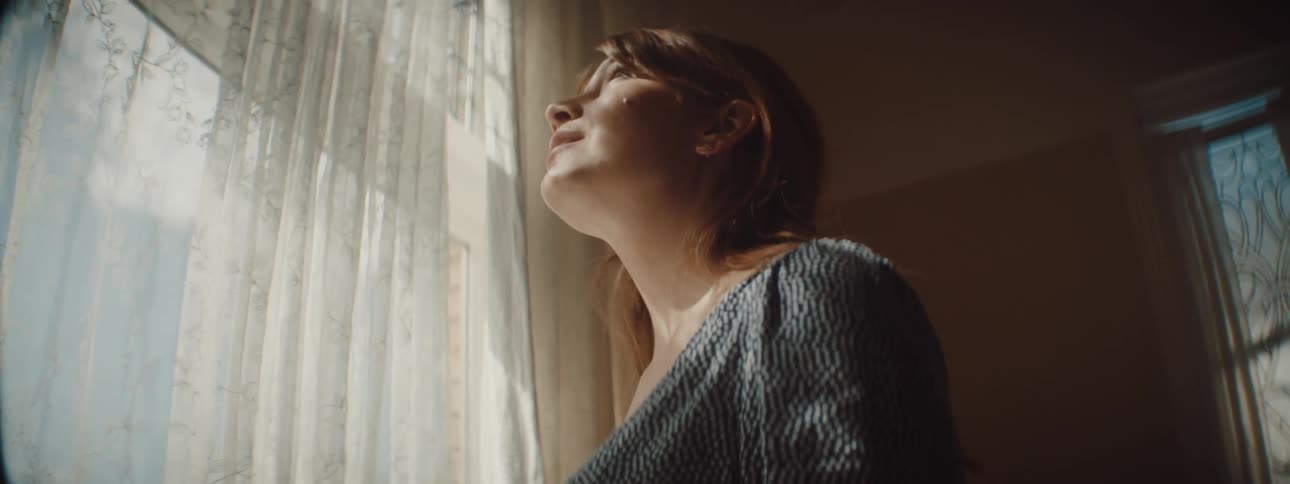 A woman in a room looking out a window with a curtain with a wall in the background