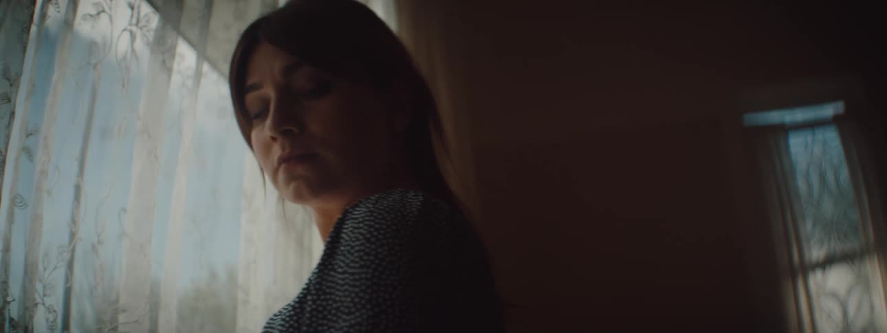 Woman with a blue dress and brown hair looks sideways in front of window in the living room at home