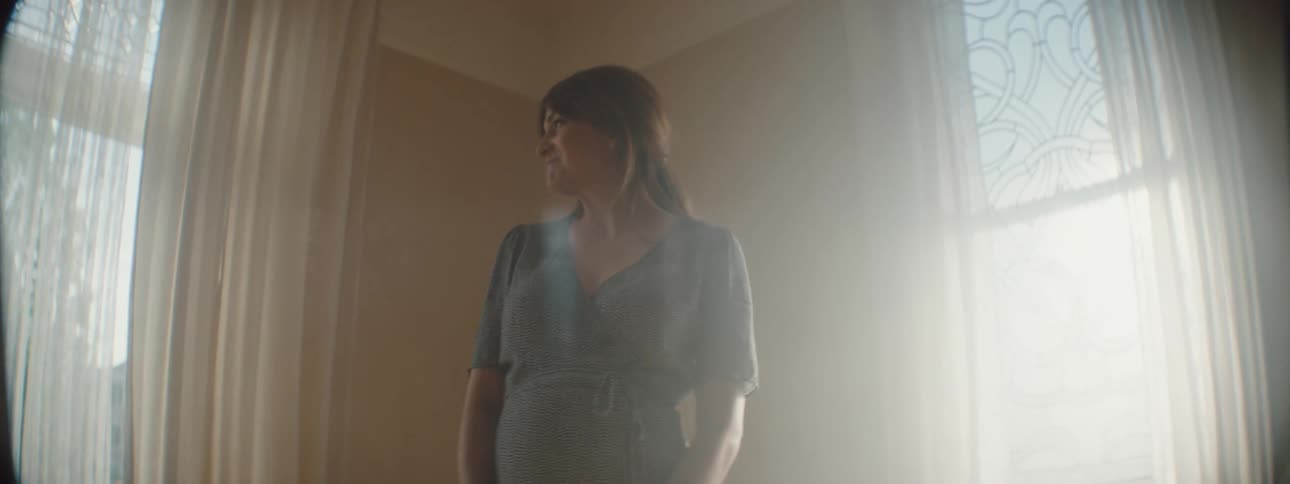 Pregnant woman in a dress smiles as she looks around a room with curtains on the windows