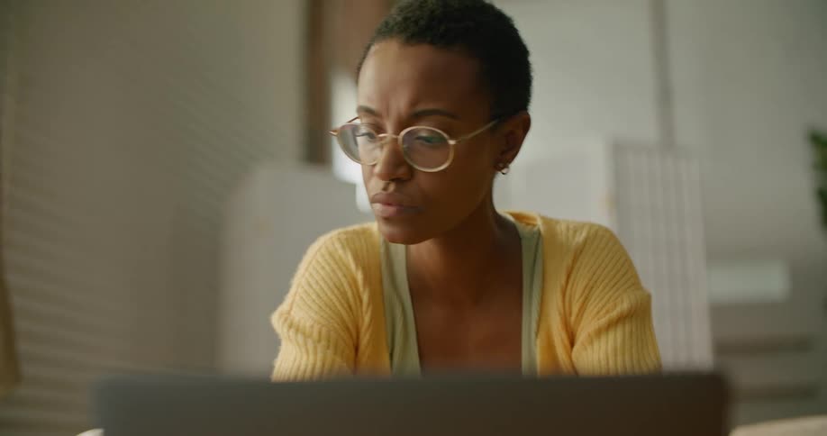 A woman with glasses using a laptop with a plant and a wall in the background