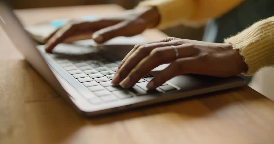 A person sitting at a table with a book and pen typing on a laptop