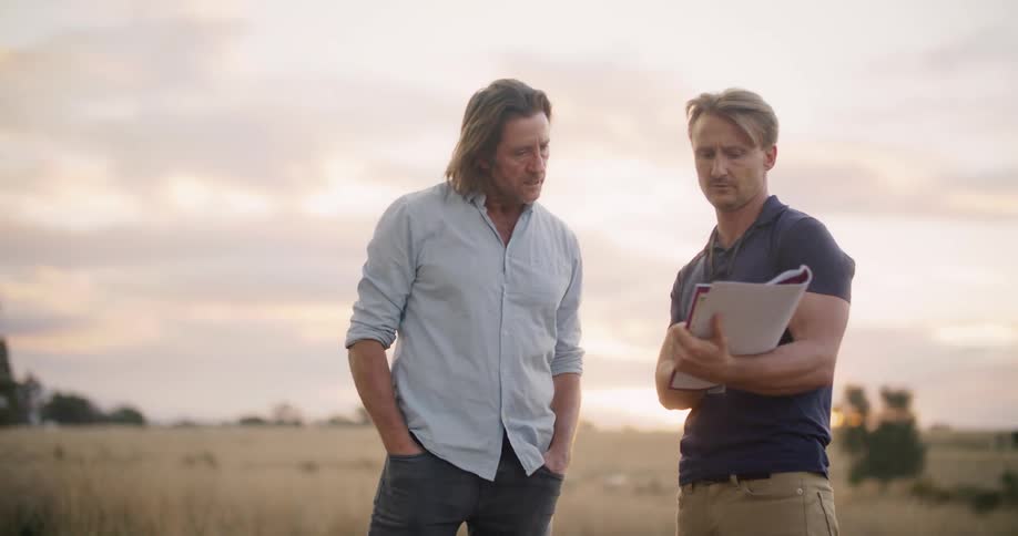 Two men stand in a field and discuss the scenery while also looking at a notebook