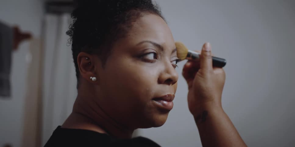 A woman smoothes her makeup with a makeup brush while standing in front of a mirror