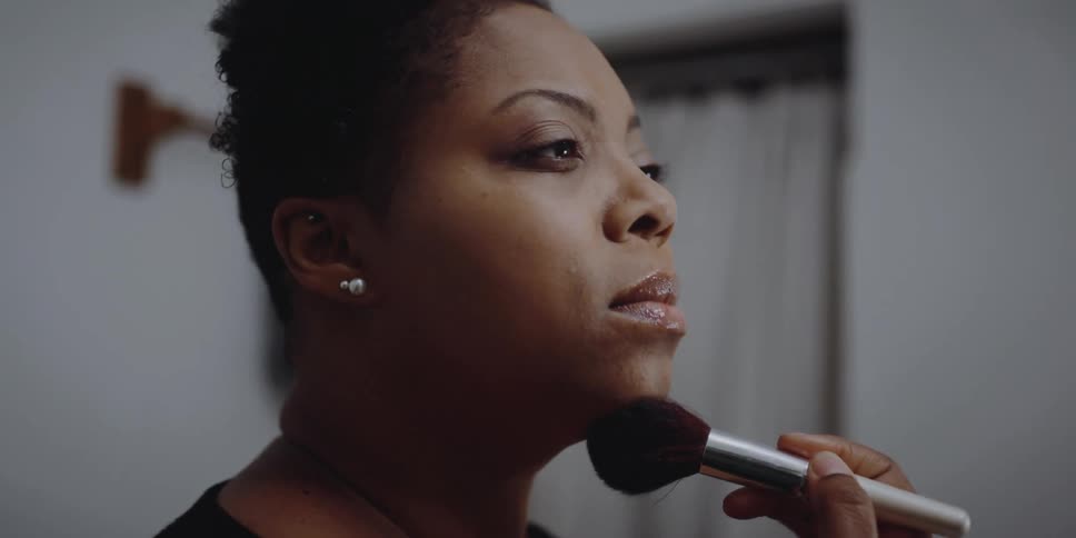 A woman in a room putting on makeup with a makeup brush with a wall in the background