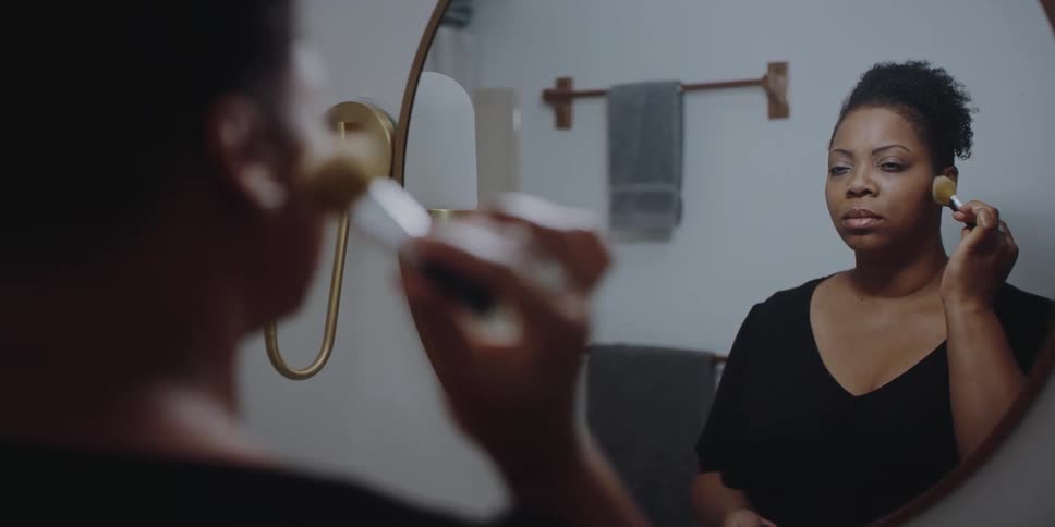 Woman stands in bathroom looking into mirror and applying foundation makeup with brush