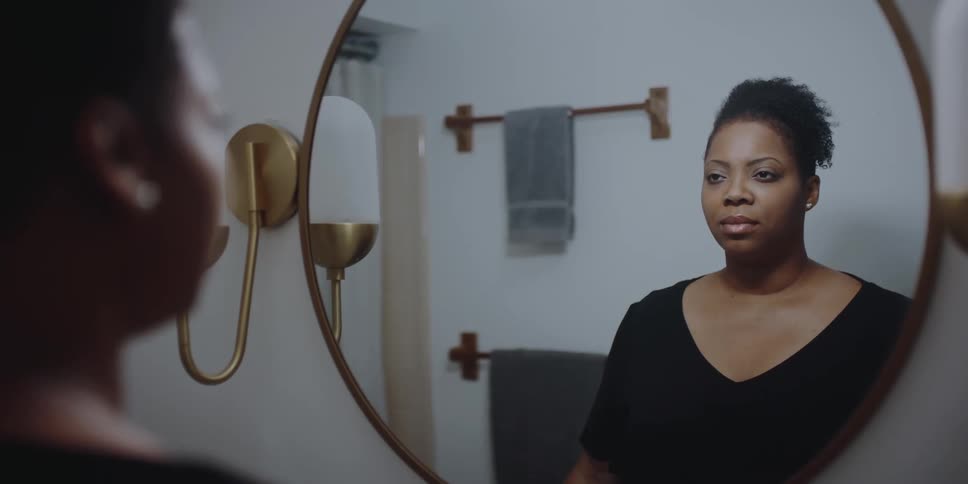 A woman stands in front of a mirror looking at her reflection with a solemn look on her face
