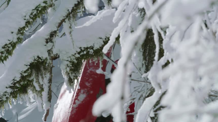 An adventurous man carries a red whitewater kayak up through snow covered trees on the shortest day of the year in heavy winter snow
