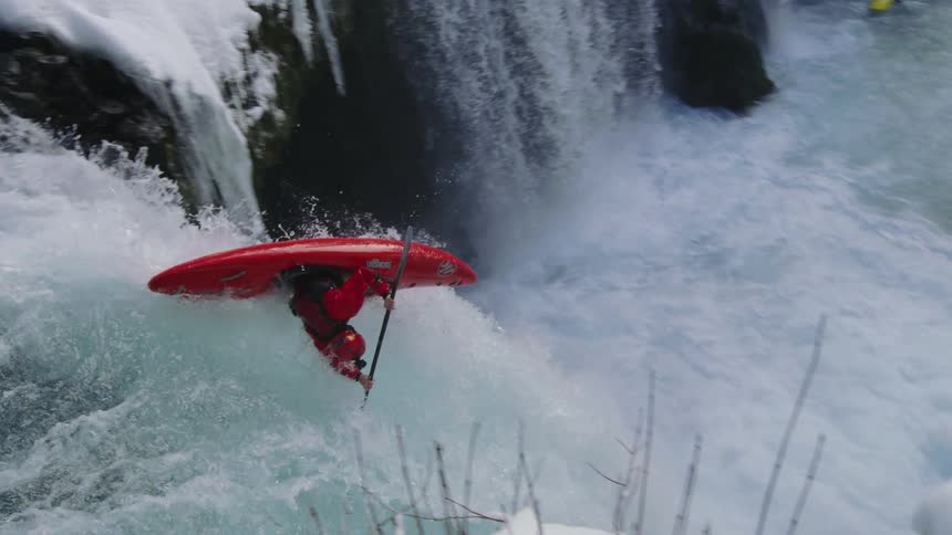 Slow motion shot of a whitewater kayak paddling up to and down a huge waterfall in the winter with a lot of snow on the ground