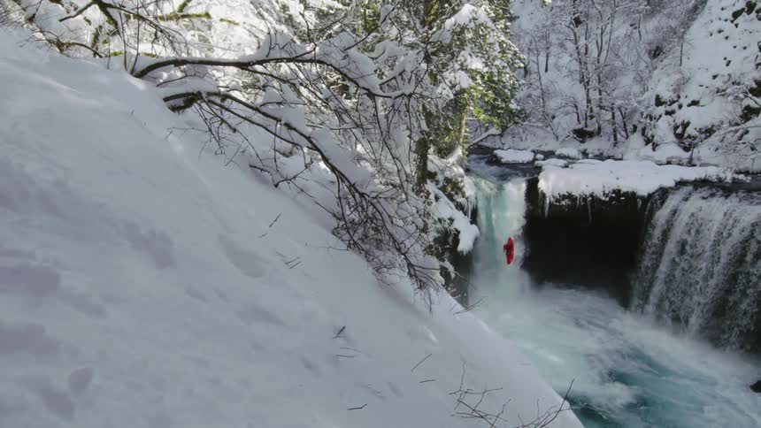 Epic shot of a rare winter snow deep in a pristine river valley, the camera pans right while a man in red kayak descends a picturesque waterfall in winter wonderland