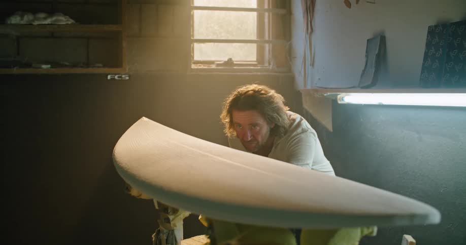 A man in a room touching and sanding a surfboard with a wall and window in the background