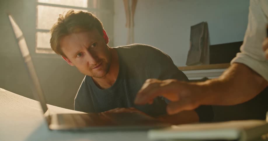 Two men in a room one using a laptop and the other one touching a surfboard with a wall and window in the background
