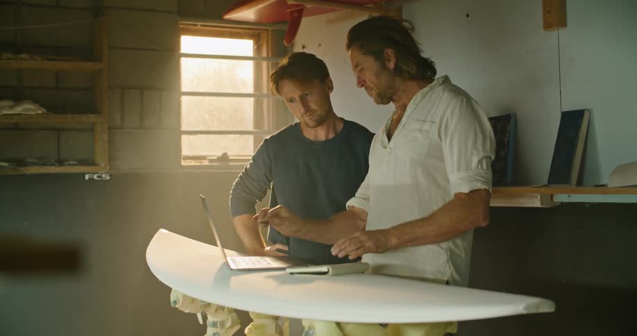 Two men in a room looking at a laptop with a wall and window in the background
