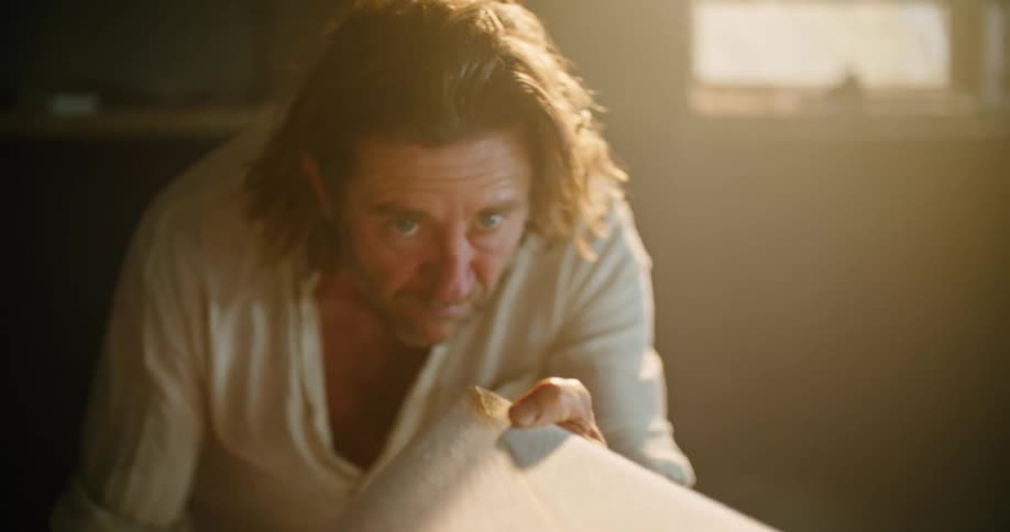 A man in a room touching a surfboard with a wall and window in the background