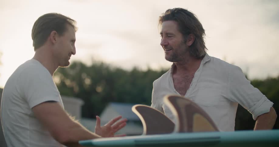 Two man excitedly discussing the design of a surfboard standing outdoors at evening