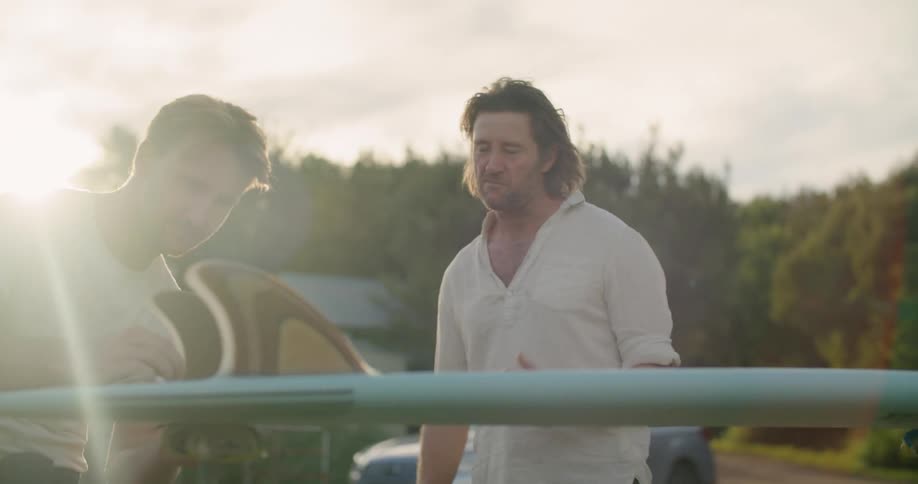 Two men stand in front of a surfboard talking to each other outside