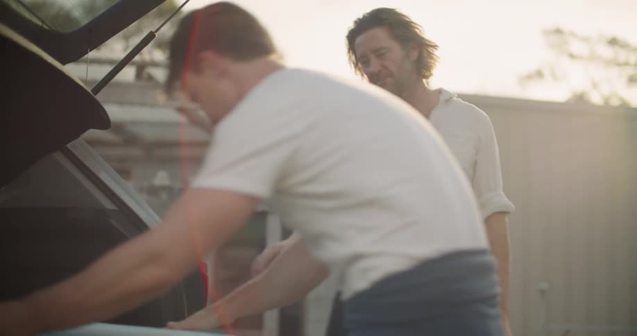 A man puts a surfboard in to the back of a vehicle and shakes another mans hand