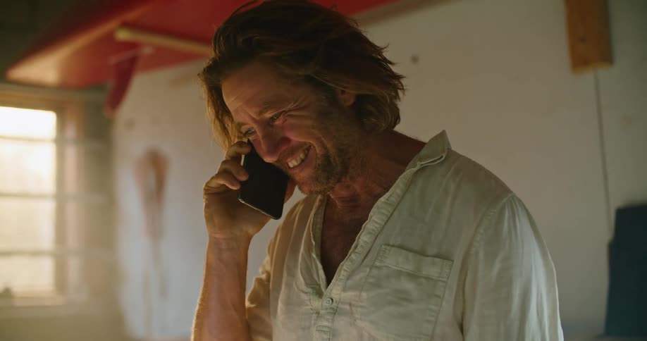 Middle aged man with long hair and a white linen shirt talks on the smartphone while using a laptop