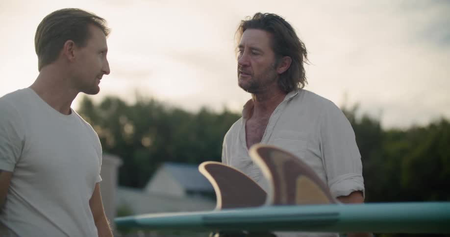 Two men standing talking and looking at a surfboard with trees the sky and clouds in the background