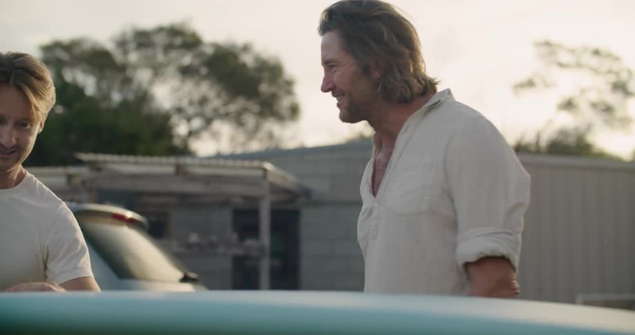 Two men standing talking and looking at a surfboard with trees the sky and clouds in the background