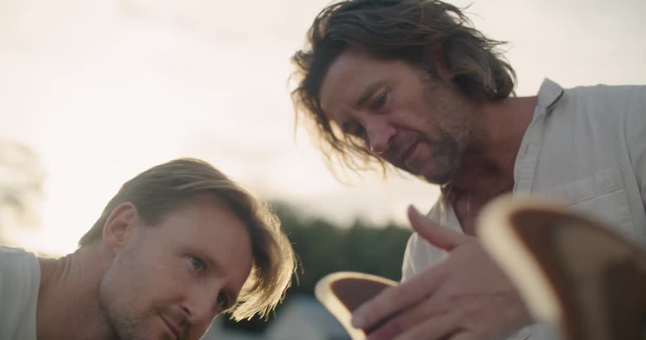 Two men discuss the wooden fins on the back of a surfboard as they stand outside