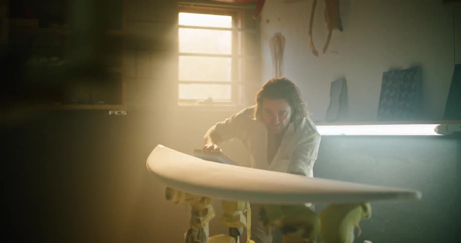 A man uses a sanding block on a surfboard in a dim workshop space