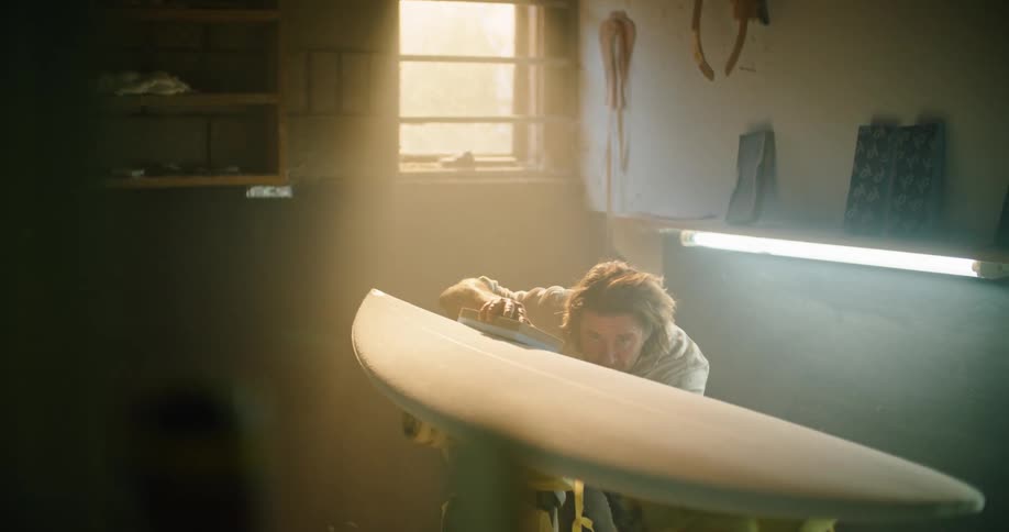 A man sands a surfboard in a small and dim workshop