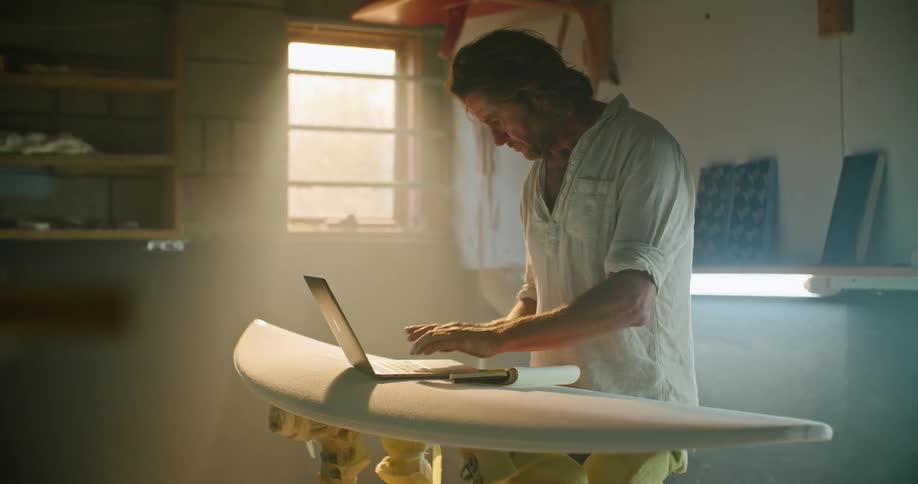 A man stands beside a surfboard in a small workshop working on a laptop that is on top of the surfboard