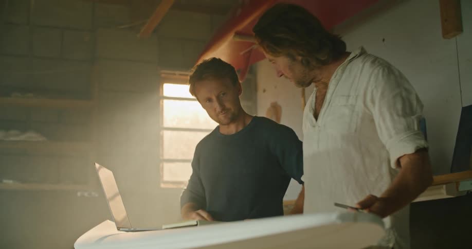 Two men talk about a surfboard laying on a stand in a small workshop