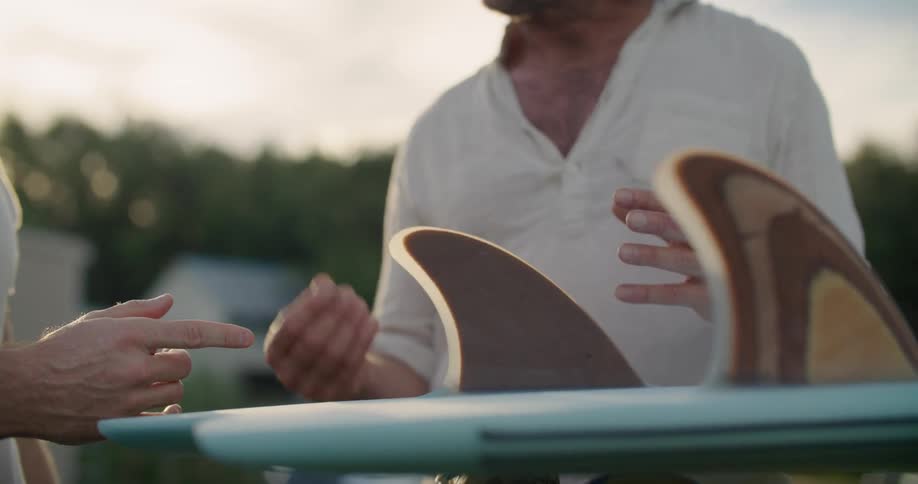 Two men talking and looking at a surfboard with trees the sky and clouds in the background