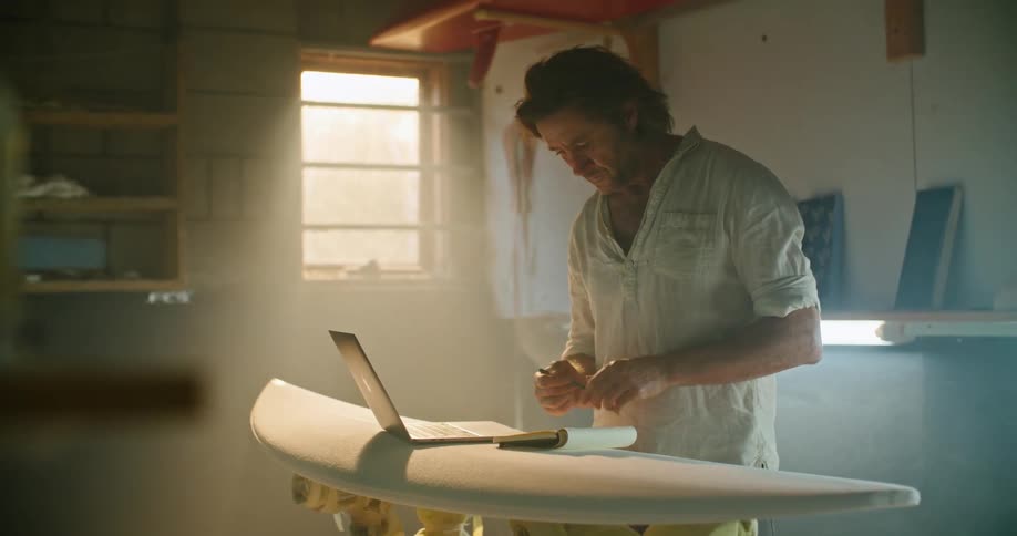 Man stands in shop typing on laptop sitting on elevated surfboard and making notes