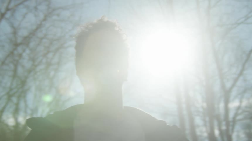 Man with a shirt and a black jacket walks in the park as the sun shines in the background