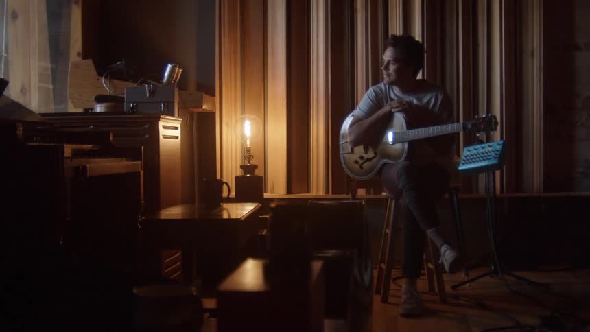 A man sitting on a stool holding a guitar inside of a room lit by a small lamp on a table