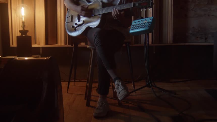 Person sits on chair in dim room lit by lamp strumming acoustic guitar