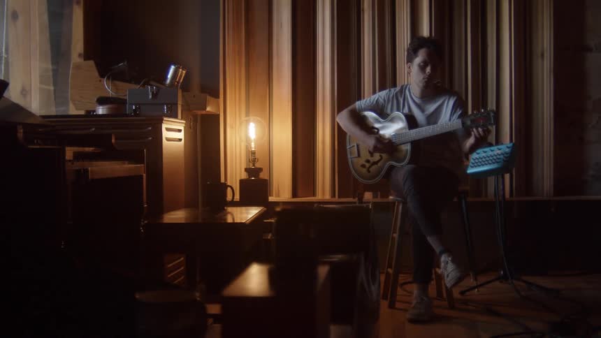 Man with a white t shirt and pants sits on a stool and makes guitar chords in the studio