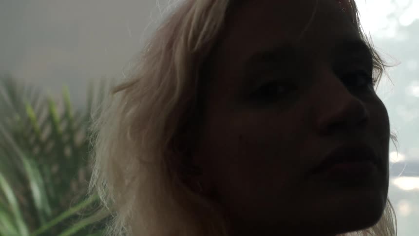 A woman in a room staring with plants a wall and a window in the background