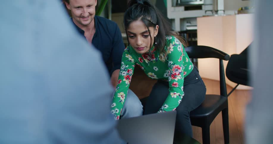 Coworkers sit around laptop on table holding meeting as one bends forward to access laptop