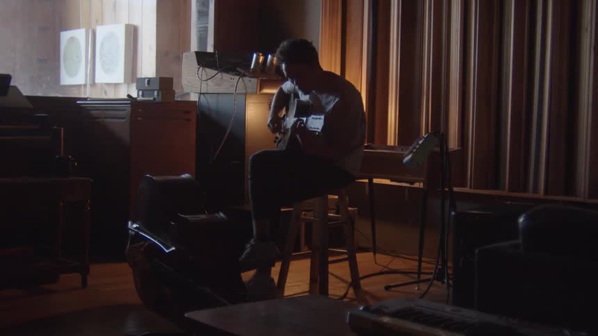 A person in a room sitting on a stool playing a guitar with a keyboard in the foreground
