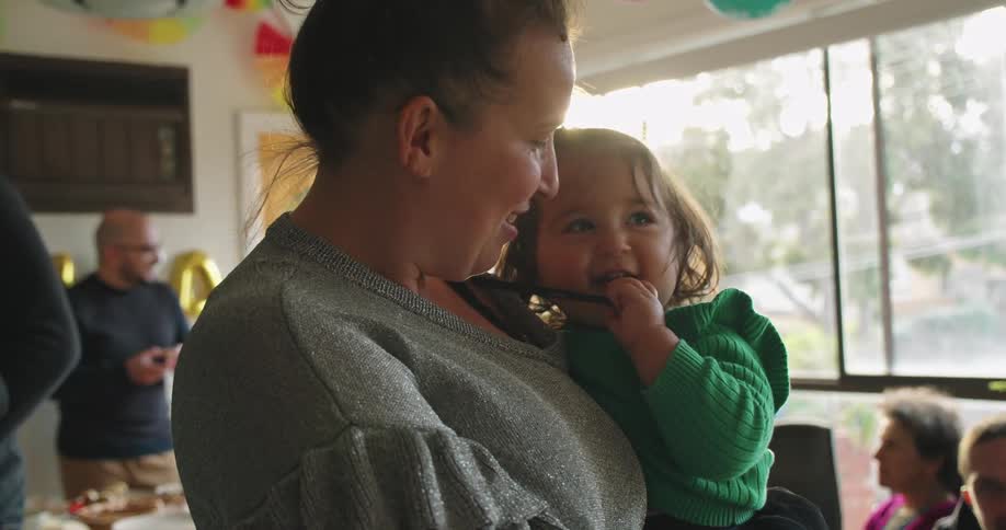 A mother holds her daughter in her arms at a family gathering indoors during the day