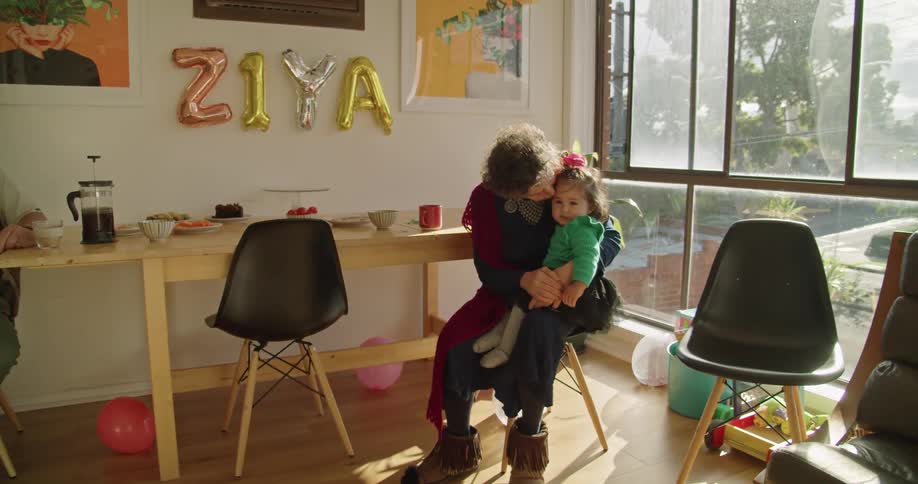 A woman sitting on a chair holding a little girl and kissing her with a wall and windows in the background