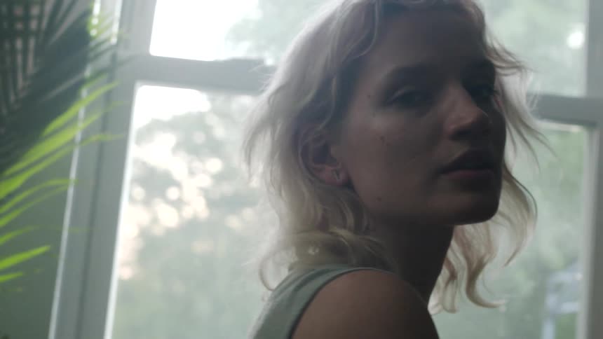 Blonde woman in a sleeveless shirt looks at the camera next to window