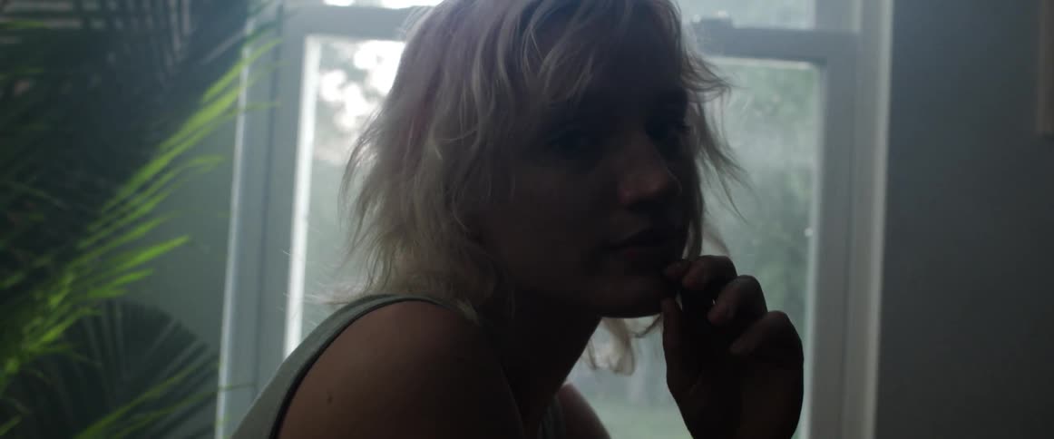 A woman in a room staring with plants a wall and a window in the background