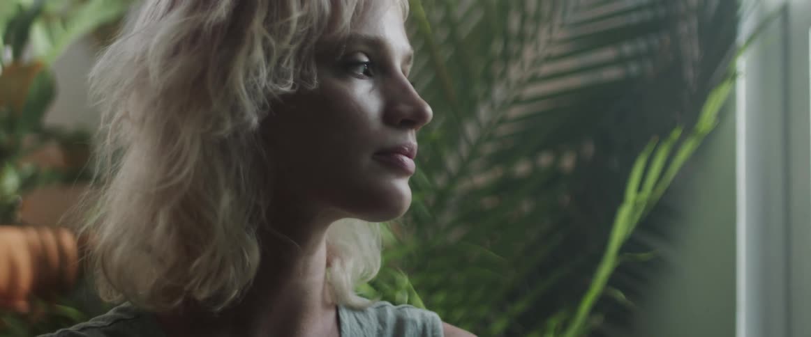 Blonde woman brings her fingers up to her chin as a green plant sits behind her