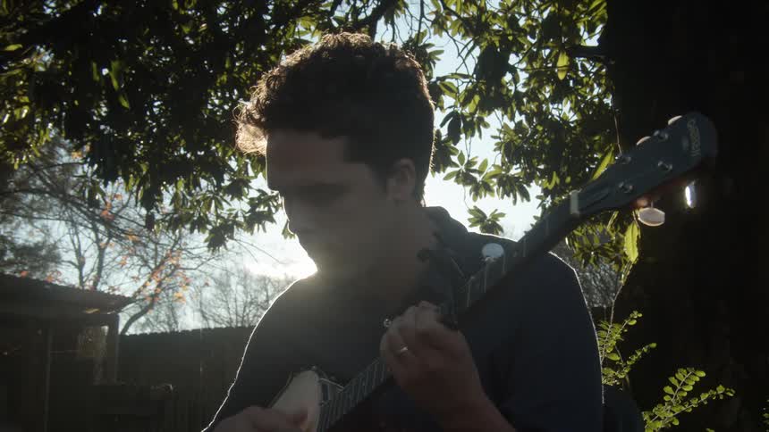 A man sitting in a backyard playing a guitar with trees the sky and clouds in the background