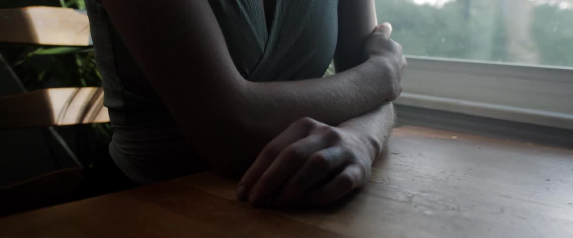 Woman sits at wooden table near window with arms crossed
