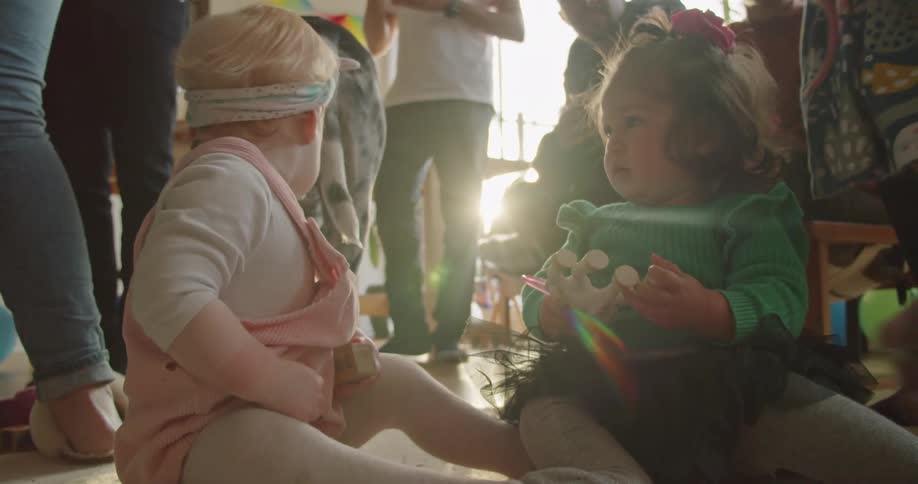 Two babies sit on the floor while adults stand nearby